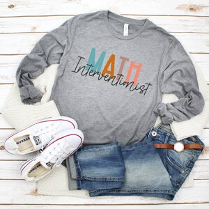 May include: A gray long-sleeved t-shirt with the text "MATH Interventionist" in colorful letters. The shirt is styled with jeans, a white cardigan, and white sneakers.