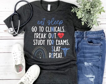 May include: A dark grey t-shirt with a white graphic print that reads "eat sleep go to clinicals freak out study for exams slay repeat". The graphic also includes a rainbow, a stethoscope, a heart, and a caduceus.