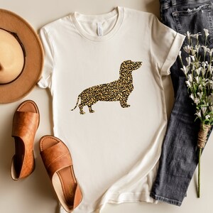 Może przedstawiać: Biały t-shirt z grafiką sylwetki  dackla w  leoparzim wzorze. Koszulka leży płasko na jasnobrązowej powierzchni z brązowym kapeluszem, brązowymi sandałami i bukietem białych kwiat&oacute;w oraz niebieskimi dżinsami.