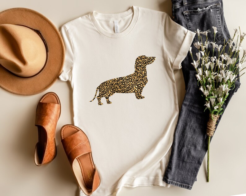 Może przedstawiać: Biały t-shirt z grafiką sylwetki  dackla w  leoparzim wzorze. Koszulka leży płasko na jasnobrązowej powierzchni z brązowym kapeluszem, brązowymi sandałami i bukietem białych kwiat&oacute;w oraz niebieskimi dżinsami.
