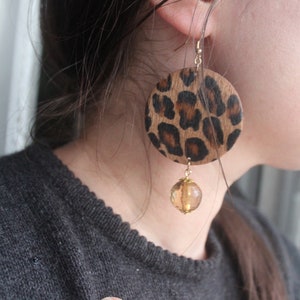 May include: A pair of gold tone earrings with a round, brown and black leopard print design and a dangling amber colored bead.