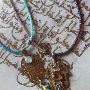 Può includere: Tre collane di perline con ciondoli a forma di mano di Hamsa dorati e un fiocco turchese. Le collane sono su uno sfondo di scrittura araba.