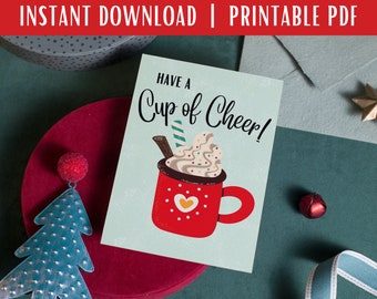 May include: A printable card with a blue background and a red mug with white whipped cream and a chocolate stick. The text on the card reads "Have a Cup of Cheer!"