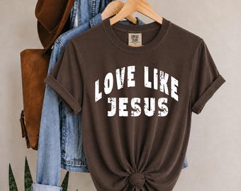 May include: A dark brown short-sleeve t-shirt with a crew neckline and rolled sleeves. The front features the white distressed text "LOVE LIKE JESUS" in a bold, arched font. The shirt has a Comfort Colors brand label inside the neck area.