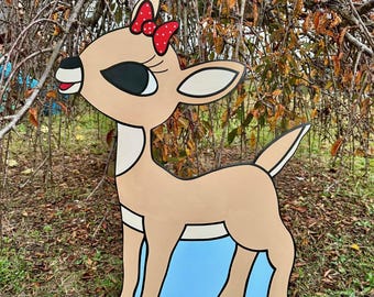 May include: A wooden cutout of a reindeer with a red bow. The reindeer has a light brown body, black eyes, and a white belly. It stands on a blue and white base, likely for outdoor display. A festive holiday decoration.