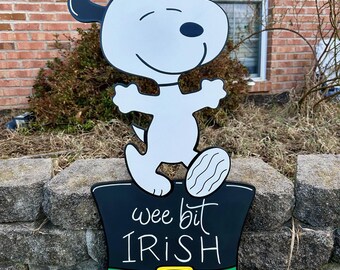 Puede incluir: Un perro de dibujos animados blanco que lleva un sombrero de copa negro con una banda verde y las palabras "wee bit IRISH" en la parte delantera. El perro está de pie sobre el sombrero con los brazos extendidos.