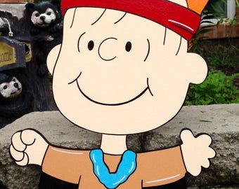 May include: Wooden cutout of a cartoon character with a red headband and a feather in the hair. The character wears a brown shirt with a blue collar and brown shoes. The cutout is designed to be placed outdoors.