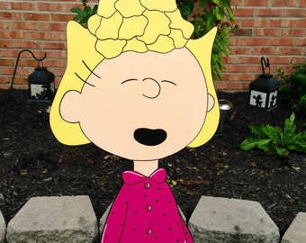 May include: A wooden cutout of a cartoon character with blonde hair and a pink dress with black dots. The character has a smiling expression and is standing on a green lawn in front of a brick wall.
