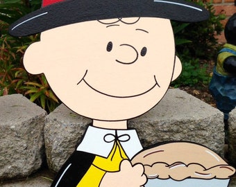 May include: A wooden cutout of a cartoon character wearing a black pilgrim hat with a red and yellow band, a yellow shirt, and a black cape. The character is holding a pie. The cutout is painted in bright colors and is suitable for outdoor decoration.