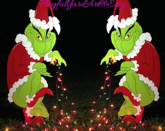 May include: Two wooden Grinch yard decorations. Each Grinch is green with a red Santa suit, hat, and boots, trimmed with white fur. They hold a string of Christmas lights. The background is dark.