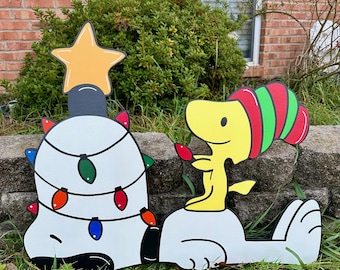 May include: Wooden Christmas yard art featuring Snoopy as a Christmas tree with a star on top and Woodstock holding a Christmas light. The figures are painted white, yellow, red, green, and black, and are designed for outdoor display.