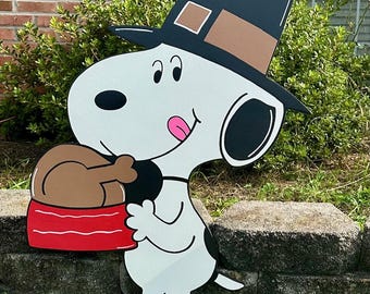 May include: A wooden cutout of Snoopy wearing a pilgrim hat, holding a turkey in a red and white bowl. The Snoopy figure is white with black ears and spots, and a pink tongue. The hat is black with a brown band.