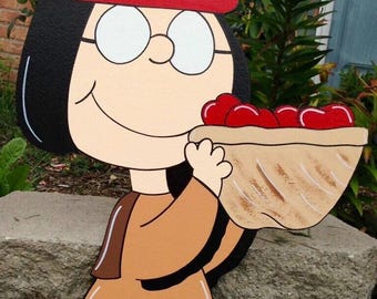 May include: A wooden cutout of a cartoon character with black hair, glasses, and a red headband, holding a basket of red cherries. The character is wearing a brown outfit with a black hem and a feather in their hair. The cutout is placed in front of a stone wall.