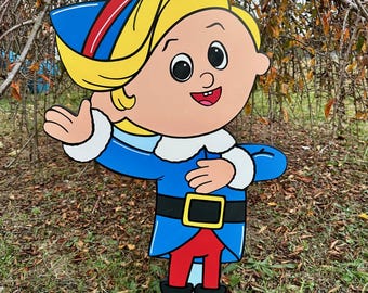 May include: A colorful, cartoonish cutout of a smiling elf with blonde hair, a blue jacket, red pants, and black shoes. The elf is wearing a blue hat with red and white stripes. The cutout is standing on green grass.