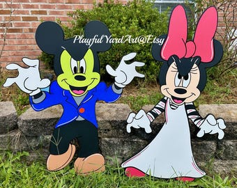 May include: Two wooden yard art figures of cartoon characters. One figure is dressed in a blue jacket and black pants, with a green face and black ears. The other figure is wearing a white dress and a pink bow. The text "Playful Yard Art @ Etsy" is visible.