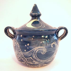 May include: A dark blue ceramic sugar bowl with two handles and a matching lid featuring a pointed finial. The exterior is decorated with white hand-drawn illustrations of swirling ocean waves, a crescent moon, and small stars across the surface.