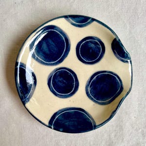 May include: A ceramic spoon rest featuring a cream-coloured base adorned with large, irregular blue polka dots. The spoon rest has a curved shape with a small lip on one side. The handmade design adds a touch of rustic charm to any kitchen.