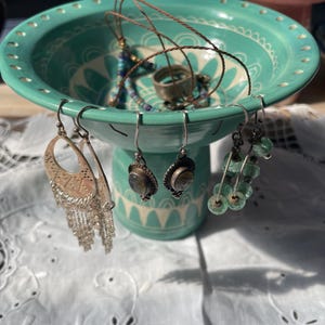 May include: A turquoise ceramic jewellery holder with a pedestal base, decorated with a white floral pattern. The holder displays various earrings, necklaces, and rings. The earrings include silver dangle earrings and beaded earrings.