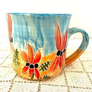 Peut inclure: Une tasse en céramique avec un motif floral peint à la main. La tasse présente un fond bleu clair avec des fleurs orange et rouges. La poignée est orange et les fleurs ont des centres noirs. La tasse mesure environ 10 cm de haut.