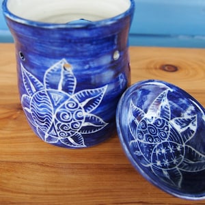 Peut inclure: Un chauffe-huile en céramique bleu et son plat assorti. L'ensemble présente un motif floral blanc peint à la main. Le chauffe-huile a une forme cylindrique avec des trous pour diffuser le parfum. Le plat est de forme ovale.