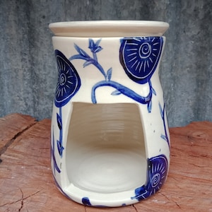 May include: A white ceramic utensil holder with a blue floral design. The holder has a rectangular opening and a wide rim. The blue design features stylized flowers and vines, adding a decorative touch to the kitchen accessory.