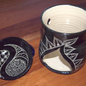 Peut inclure: Un chauffe-huile en céramique noire avec un couvercle. Le chauffe-huile présente un motif de feuilles blanches et une découpe pour une bougie. Le couvercle présente un motif cachemire blanc. L'intérieur est de couleur crème.