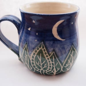 May include: Ceramic mug with a night sky design. The mug features a dark blue background with a crescent moon, stars, and green leaf-shaped patterns. The handle and rim are also blue.