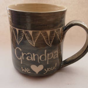 May include: A handmade ceramic mug with a dark blue-grey glaze. The mug features the word "Grandpa" in white script, along with a white heart and the words "we you!" A white geometric design encircles the top.