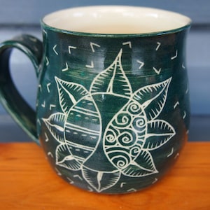May include: A handmade ceramic mug with a dark teal glaze and a white floral design. The mug features a large handle and a cream-colored interior. The floral design includes detailed leaves and spiral patterns.