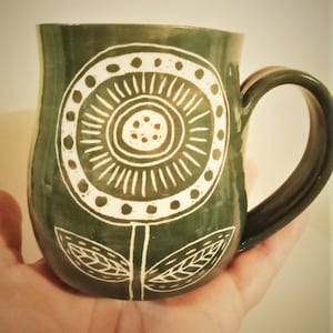 May include: A dark green ceramic mug with a white floral design. The design features a large flower with a circular centre and radiating lines, along with leaves on the stem. The mug has a curved handle.