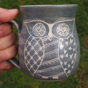 May include: A blue-grey ceramic mug with a white owl design. The owl is decorated with geometric patterns and hearts. The mug has a curved handle and is held in a hand. The mug is suitable for hot drinks.