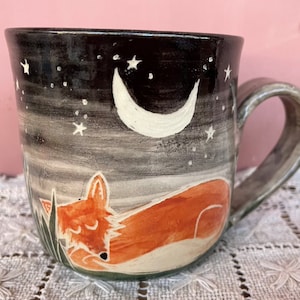 May include: A ceramic mug with a hand-painted design. The mug features a sleeping orange fox under a crescent moon and stars against a night sky background. The handle and rim are grey.