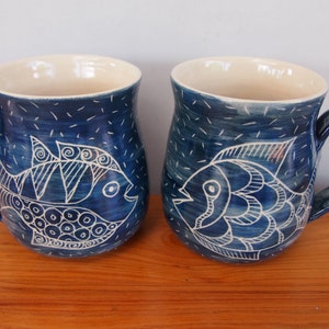 May include: Two ceramic mugs with a blue glaze and white fish designs. The mugs have a curved handle and a cream-colored interior. The fish are hand-drawn with intricate details. The mugs are approximately 4 inches tall.