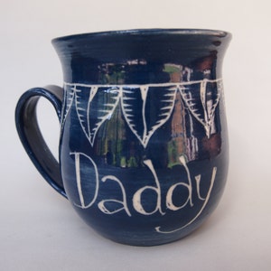 May include: A navy blue ceramic mug with a curved handle. The mug features the word "Daddy" in white script, along with a white leaf design. The mug is a unique gift idea.