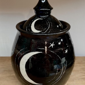 May include: Black ceramic jar with a lid, decorated with white crescent moons and stars. The jar has a rounded body and a conical lid. The design is hand-painted, giving it a unique, artistic look. The jar is likely used for storage.