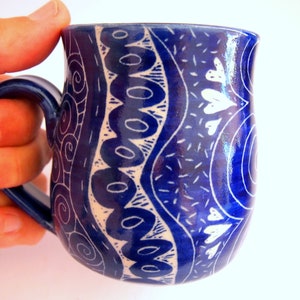May include: A blue ceramic mug with a curved handle, decorated with white patterns. The patterns include swirls, circles, and heart shapes. The mug is held by a hand.
