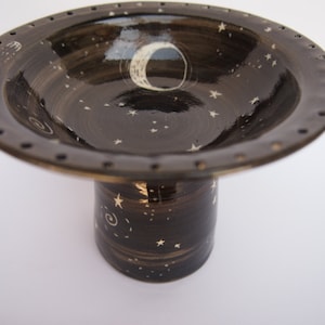May include: A ceramic pedestal bowl with a dark brown glaze, decorated with white stars and a crescent moon. The rim features small, evenly spaced holes. The bowl is elevated on a cylindrical base, also adorned with stars.
