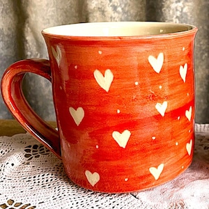 May include: A handmade ceramic mug with a red exterior decorated with white heart shapes and small white dots. The mug has a large handle and a cream-coloured interior. The mug is on a white lace doily.