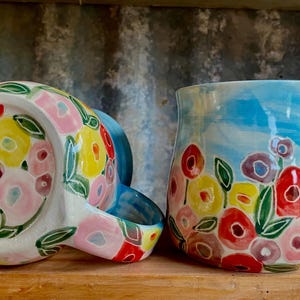 May include: A ceramic jug and a spoon rest, both decorated with a colourful floral design. The jug is light blue with red, yellow, and purple flowers. The spoon rest is white with a matching pattern. Hand-painted.
