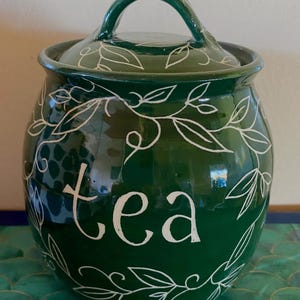 May include: A dark green ceramic tea canister with a lid and handle. The word "tea" is written in white script on the front, surrounded by white leaf and vine designs. The canister is a rounded shape.