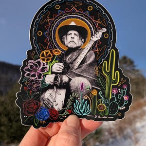 May include: A black sticker featuring a black and white image of a musician with a guitar, surrounded by colorful flowers and cacti. The sticker has a decorative border and the artist's signature, "D. Rosalee."
