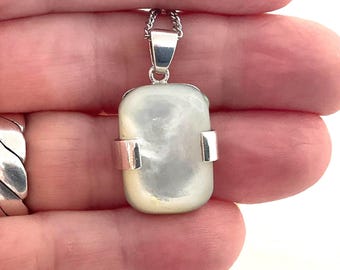 May include: A rectangular pendant with a pearlescent, off-white stone is suspended from a silver chain. The stone is encased in a silver setting with two silver bands. The pendant is held by a hand.