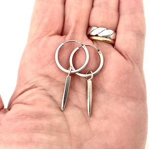 May include: A pair of silver hoop earrings with a dangling, elongated, rectangular charm. The earrings are displayed against a light background, showcasing their minimalist design and metallic sheen. The hoops are approximately 2.5 cm in diameter.