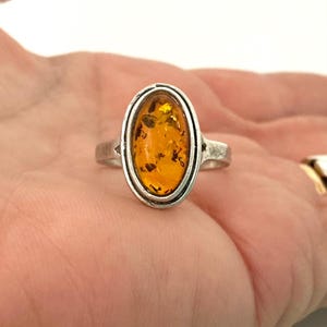 Puede incluir: Anillo de plata con una piedra de ámbar ovalada con inclusiones oscuras. El ámbar está engastado en un bisel de plata con un borde doble. El anillo se muestra en una mano.