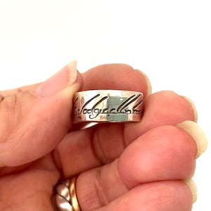 May include: A wide silver band ring featuring a polished metallic finish and a dark, engraved cursive script wrapping around the exterior surface. The ring has a flat profile and a smooth, reflective texture across the visible metal band.