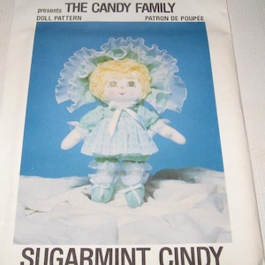 May include: A vintage dollcraft sewing pattern for a doll named "Sugarmint Cindy" from "The Candy Family". The pattern features a doll with blonde hair, a blue and white dress, and a white bonnet.