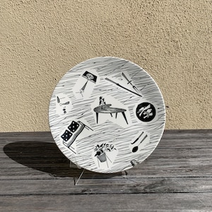 May include: A black and white ceramic plate with a mid-century modern design featuring furniture, plants, and household items. The plate has a white background with black line drawings.