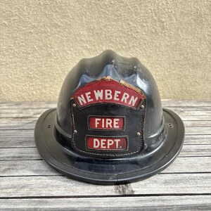 May include: A black fire helmet featuring a front leather shield with red and white text that reads NEWBERN FIRE DEPT. The helmet has a curved brim and a molded crown, showing signs of wear on the leather patch and the helmet surface.