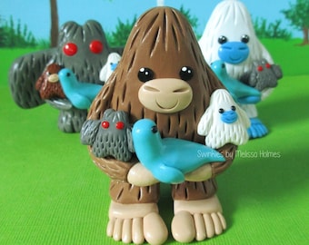 May include: Three handmade polymer clay figurines of mythical creatures. A brown Bigfoot holds a blue bird and a gray bird. A white Yeti holds a blue bird and a gray bird. A gray creature with red eyes holds a brown bird and a blue bird. The figurines are on a green surface. "Swinkies by Melissa Holmes" is written on the surface.