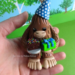 May include: A brown Bigfoot figurine wearing a blue party hat with white polka dots. The figurine holds a small chocolate cake with colorful sprinkles and a green gift box with a blue bow. The text "Swinkies by Melissa Holmes" is visible.
